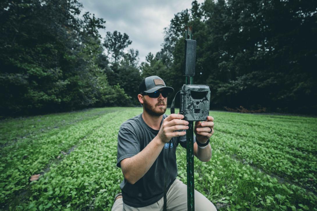 Man attaching Spartan GoLive trail camera to a post in a field surrounded by trees.